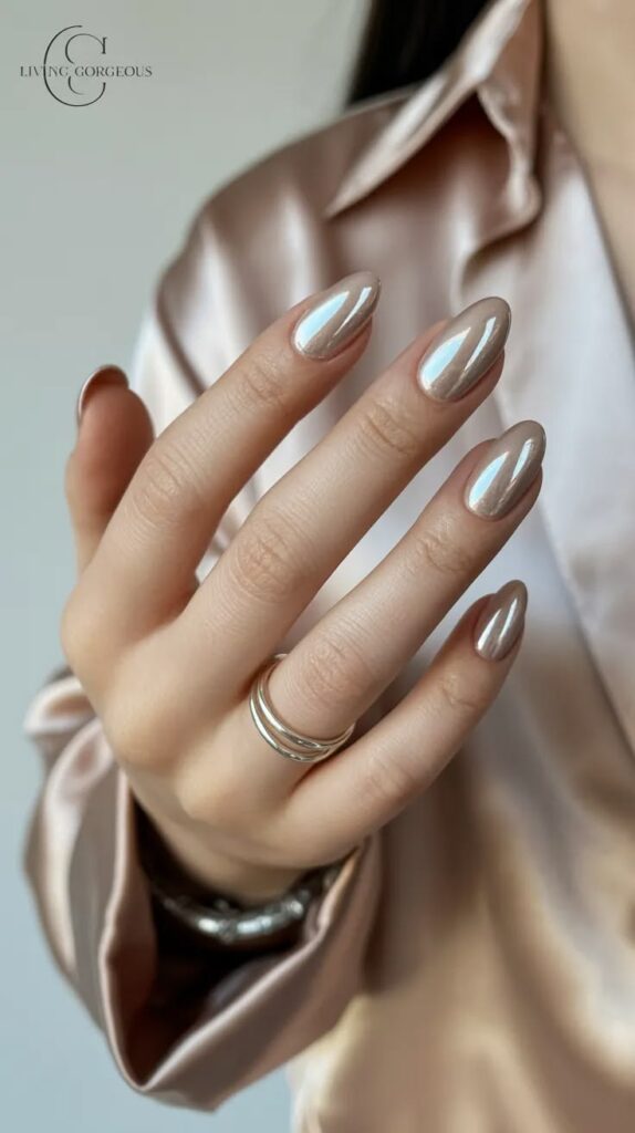 : A hand with long, elegant almond nails painted in a luminous, metallic champagne silver chrome polish, styled with a double-band silver ring and a champagne satin long-sleeve blouse.