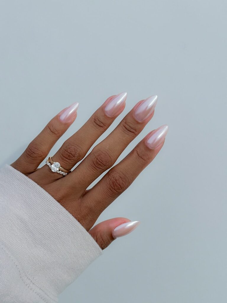 Against a light background, a manicured hand with an oval-diamond ring displays long, sharp-oval wedding nails polished with a glossy, sheer pink pearlescent shimmer.
