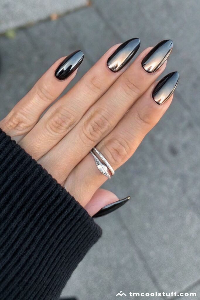 Long almond nails with a dark, iridescent black and gold metallic polish against a black ribbed sleeve.