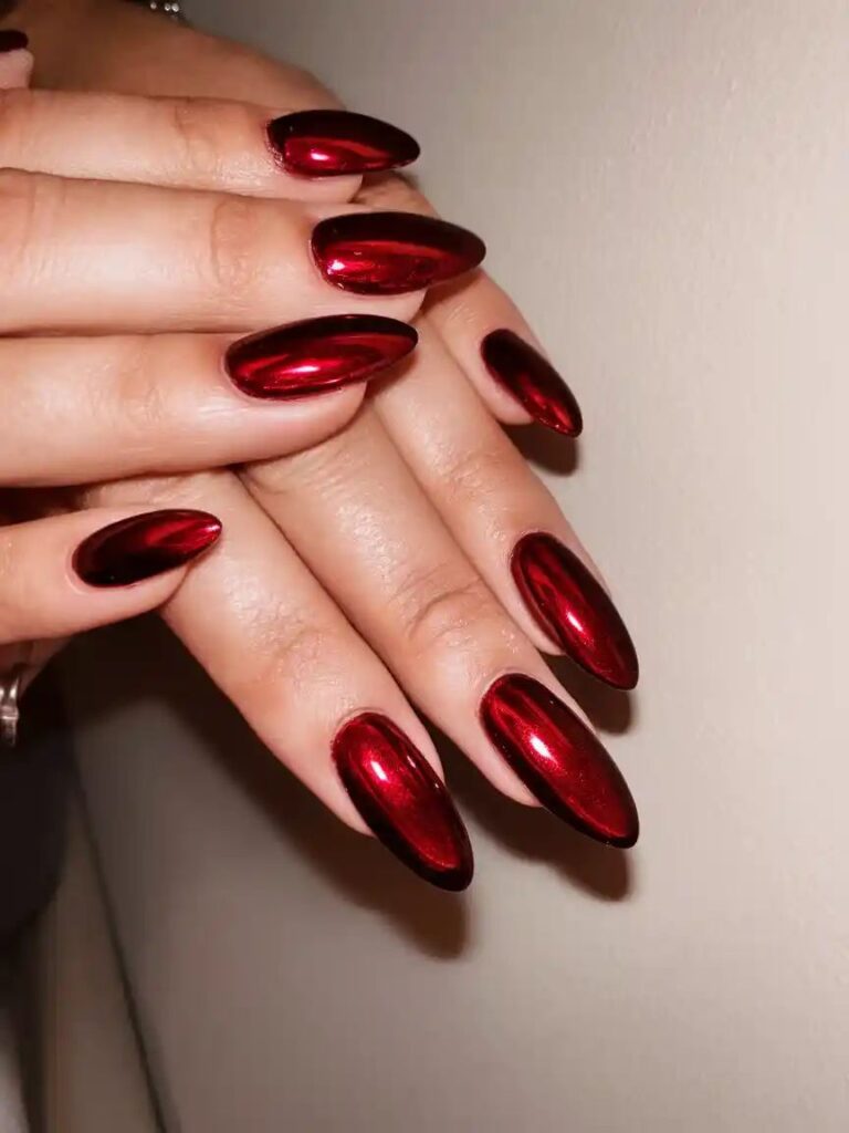 A stunning top-down view of two hands resting over each other with long, pointed almond nails coated in a rich, metallic dark ruby red chrome polish with bright crimson reflections.