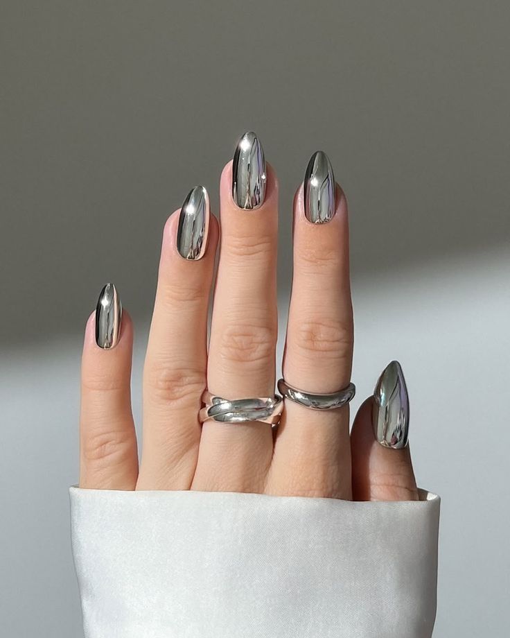 A close-up of a hand with long, mirror-finish silver metallic nails and thick silver rings.