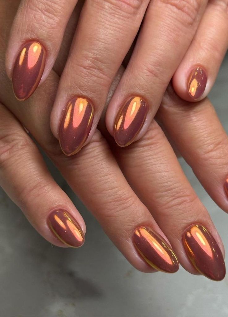 A close-up of hands with neatly shaped almond nails featuring a luminous, metallic warm bronze chrome finish that reflects soft orange and peach undertones.