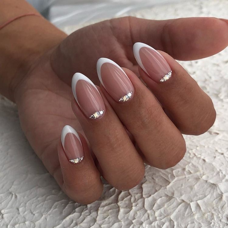 A close-up of a hand showcasing long, almond-shaped French bridal nails with bright white tips and a warm nude base, each highlighted by three shimmering rhinestones nestled along the cuticle line.