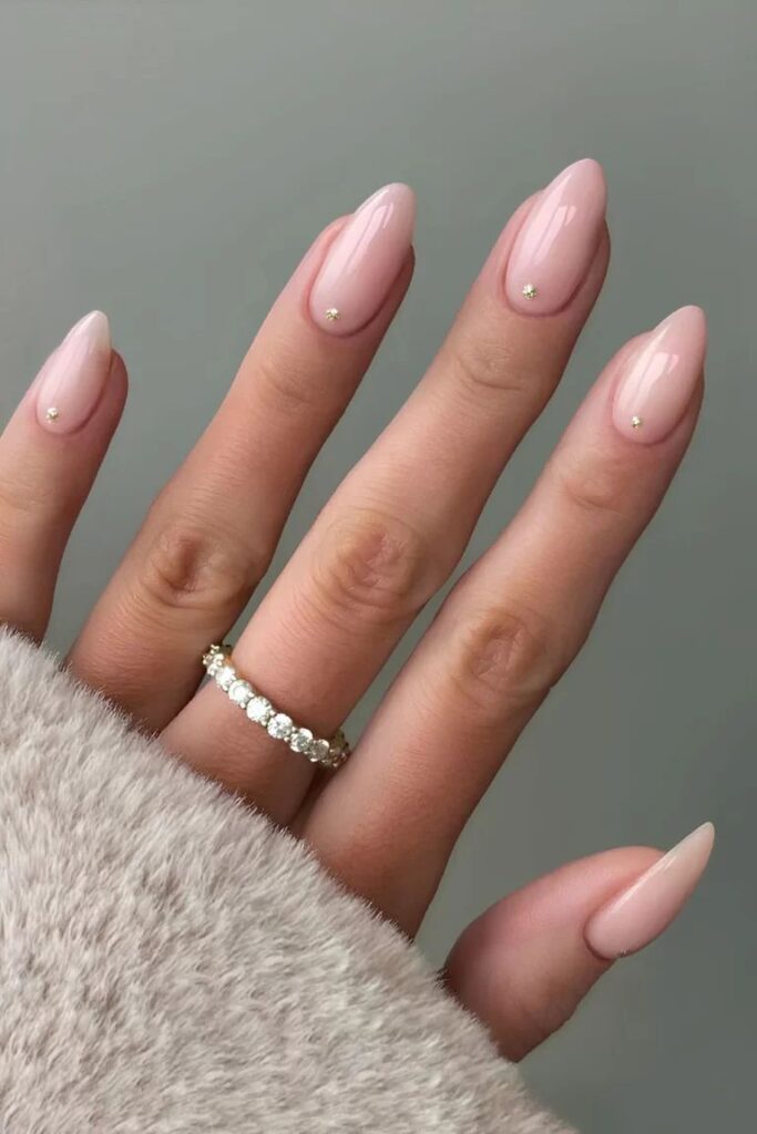 A high-end editorial shot of a hand wearing a diamond eternity band, showcasing long almond wedding day nails for bride in a sheer, high-shine nude pink with tiny gold pinprick studs at the cuticles.