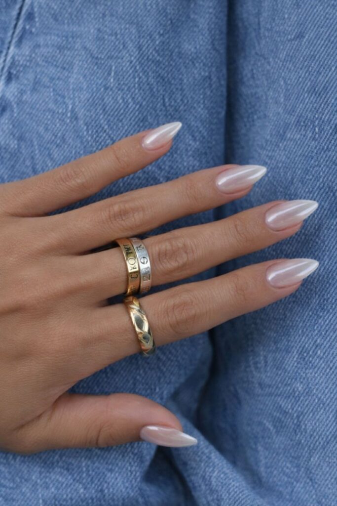 A well-manicured hand against blue denim has sharp, oval-shaped wedding day nails polished with a glossy, pearlescent pearl glaze, and features two gold rings.