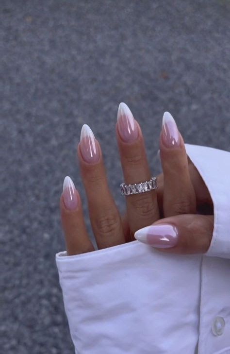 A hand with a white cuff has long, almond-shaped French nails coated in a shimmering, opalescent chrome finish, wearing an emerald-cut eternity band.