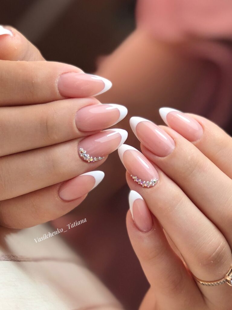 Two hands are brought together to display a pristine French manicure on almond nails, with a delicate crescent of tiny crystals accenting the base of the ring fingers.
