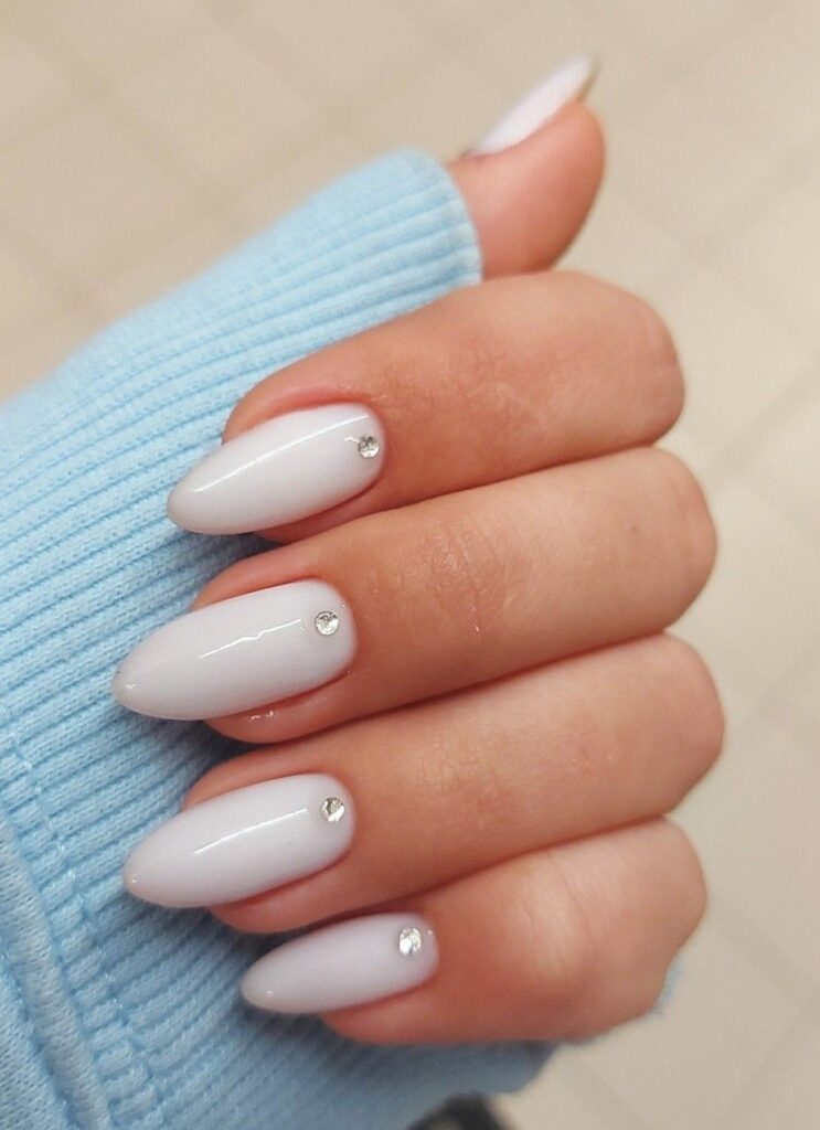 A well-groomed hand with long, oval milk-white bridal nails, where each nail is precisely adorned with a single, small silver rhinestone centered right above the cuticle.