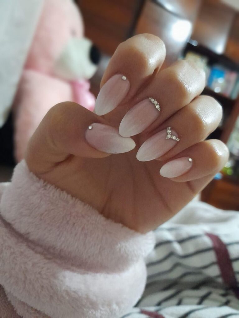 A hand showcasing a soft white-and-pink ombré manicure on sharp almond nails, with various fingers creatively adorned with single rhinestones, a crystal cuff, and a chevron rhinestone design.