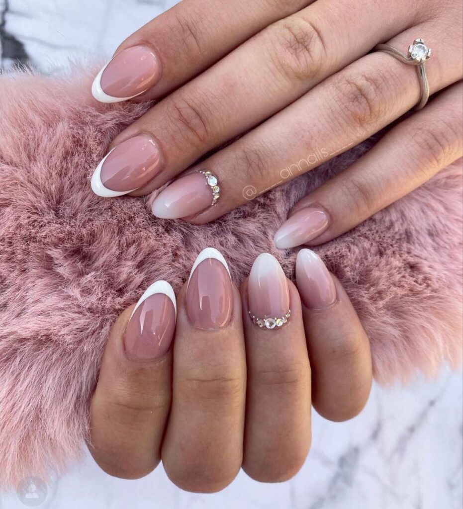Two hands resting on pink faux fur showcasing a mix of French tips and soft pink-to-white ombré wedding day nails for bride, highlighted by a curved line of rhinestones at the cuticle base of the ring fingers.