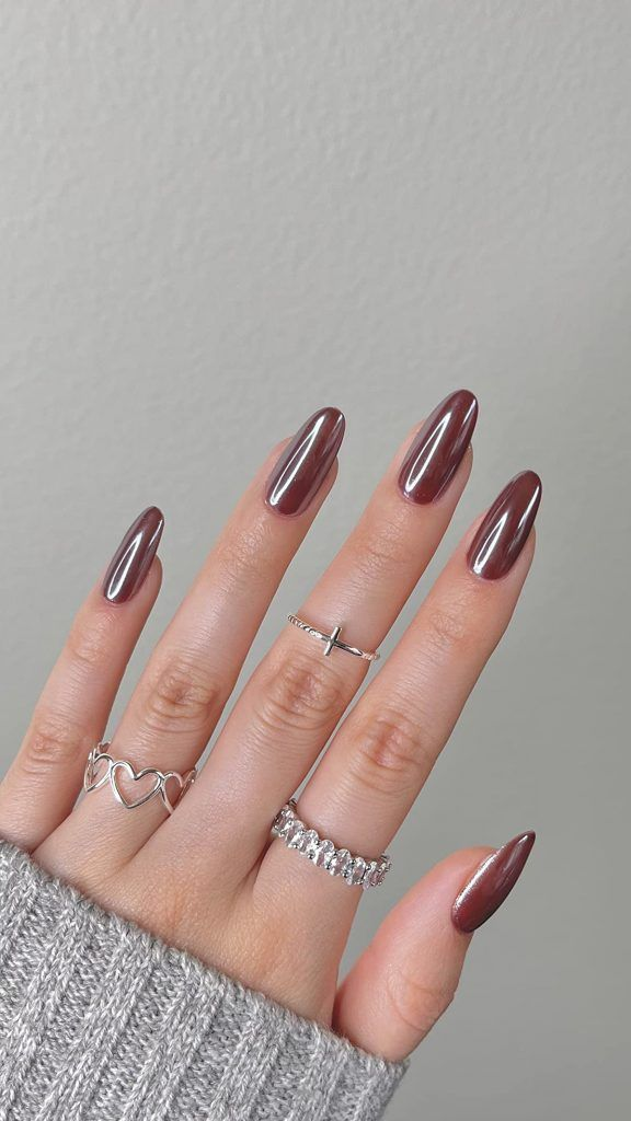 A hand with long almond-shaped nails painted in a metallic mocha-brown chrome finish. The hand is styled with three silver rings—one with a heart pattern, one with a small cross, and one eternity band—resting against a gray knit sweater.