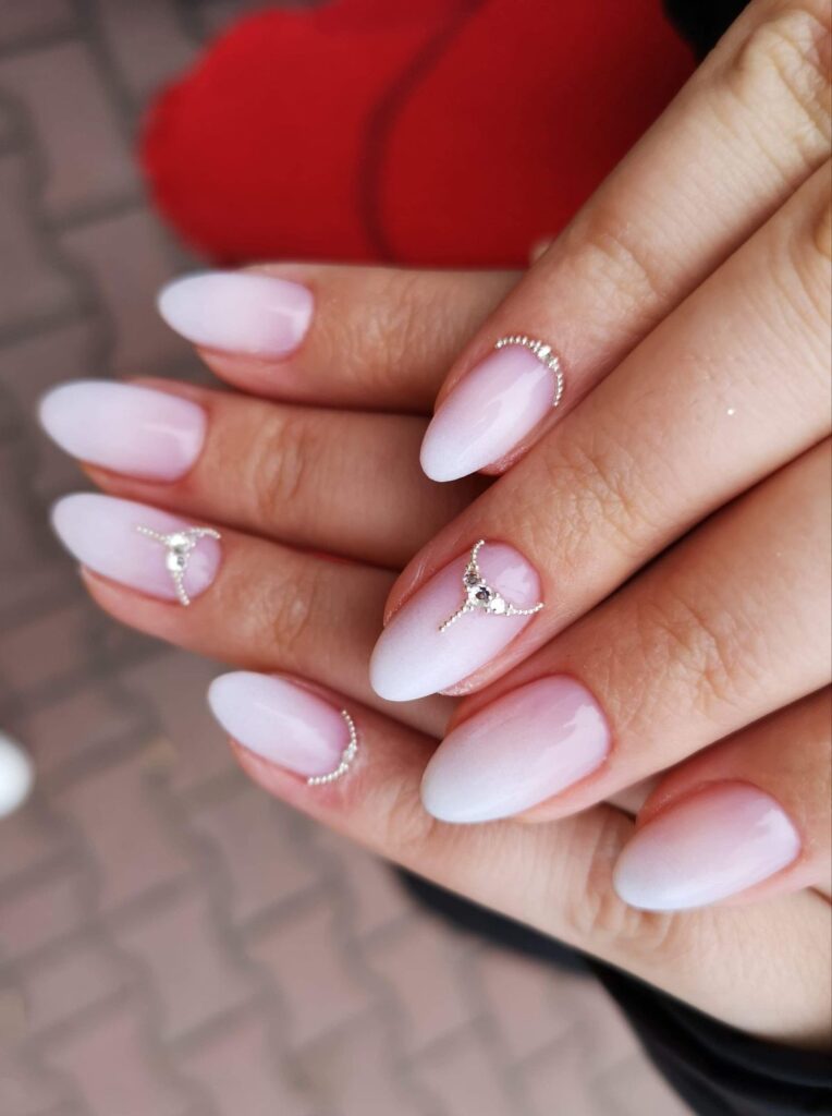 A close-up view of hands featuring oval-shaped, soft pink-and-white ombré nails accented by delicate, tiara-style curved silver crystal embellishments at the cuticles.