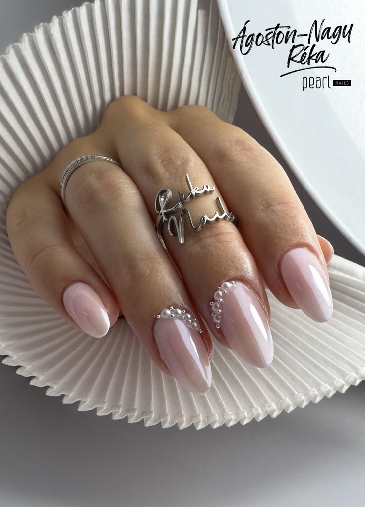 A close-up look at a hand displaying long, almond-shaped wedding day nails for bride polished in a sheer, milky pink with a delicate, sweeping arc of white pearls gracing the side cuticles.