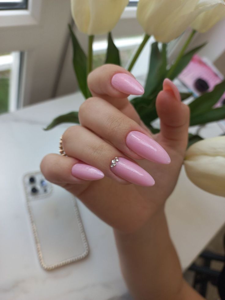 A hand with sharp, almond-shaped classy nails coated in a shimmering, pale bubblegum pink polish, accented by three tiny rhinestones nestled near the cuticle of the ring finger.