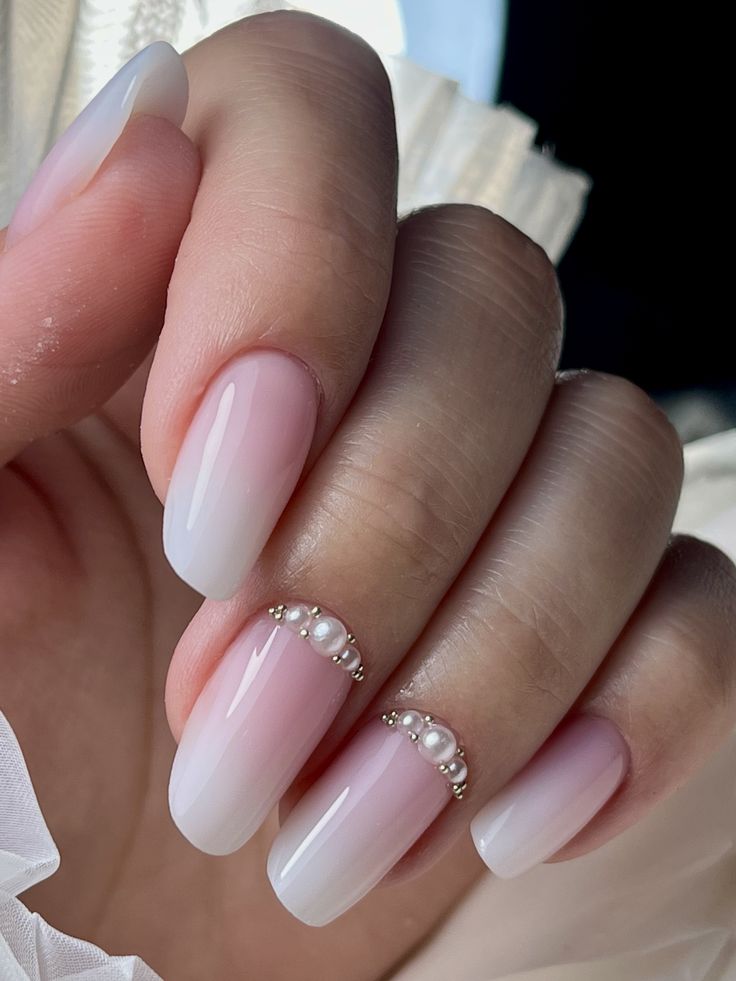 A manicured hand resting on a pleated white surface shows off glossy, high-shine soft pink wedding nails with an elegant side-swept accent of white pearls and silver microbeads.