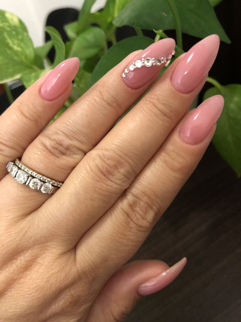 Long, elegant almond bridal nails in a glossy mauve-pink hue, highlighted by a graceful curve of graduating clear rhinestones along the side of the ring finger.
