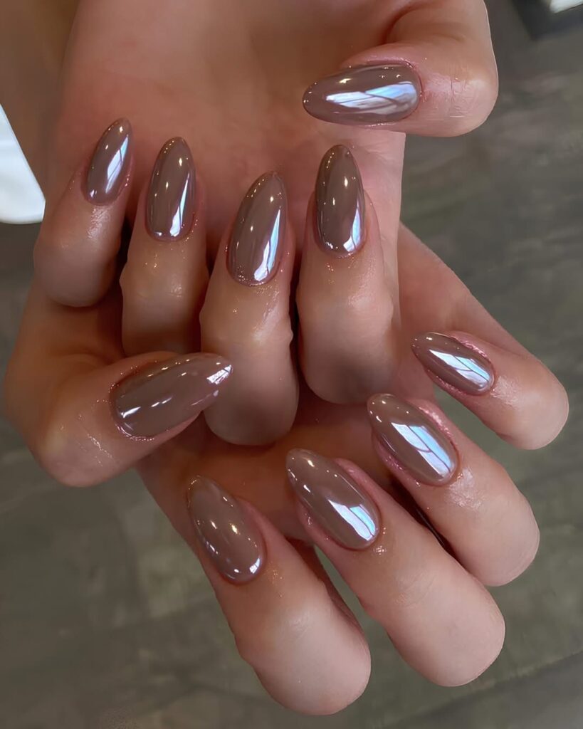 A hand with almond-shaped nails in a warm, shimmering chestnut brown metallic polish against a dark wood background.