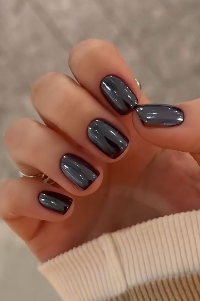 A close-up of a hand showing short, rounded square nails with a highly reflective, dark gunmetal gray chrome finish, styled with a beige ribbed sweater cuff.