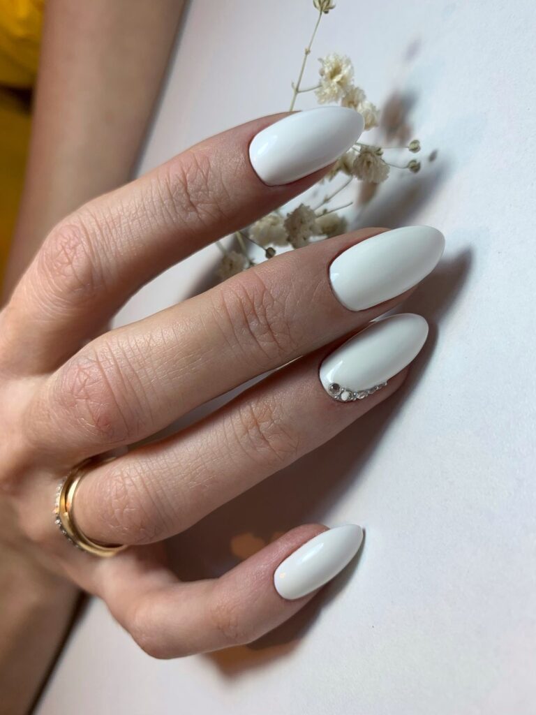 A hand rests next to white baby's breath flowers, showing off long, opaque white bridal nails with a small, curved line of crystals at the base of the ring finger.