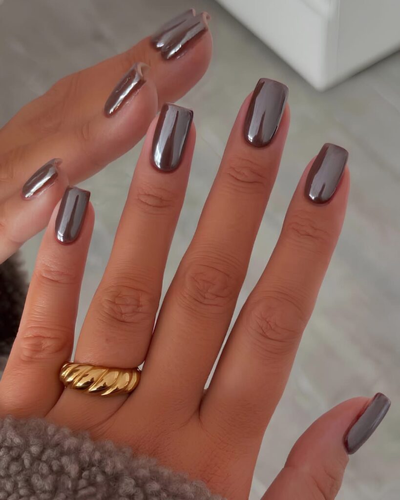 A hand showcasing medium-length square nails coated in a rich, metallic espresso brown chrome finish, accessorized with a textured gold ring and a fuzzy brown sweater.