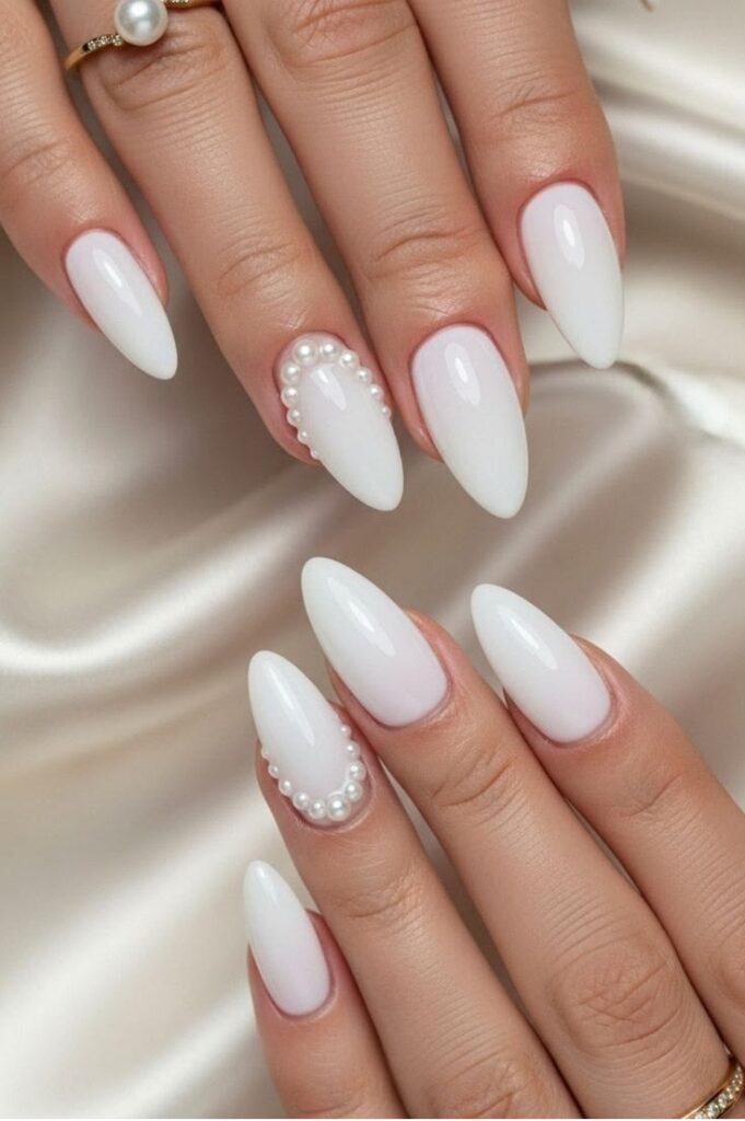 Against a cream satin background, two hands display stark white, almond-shaped wedding nails, featuring a single accent nail fully outlined in miniature white pearls.