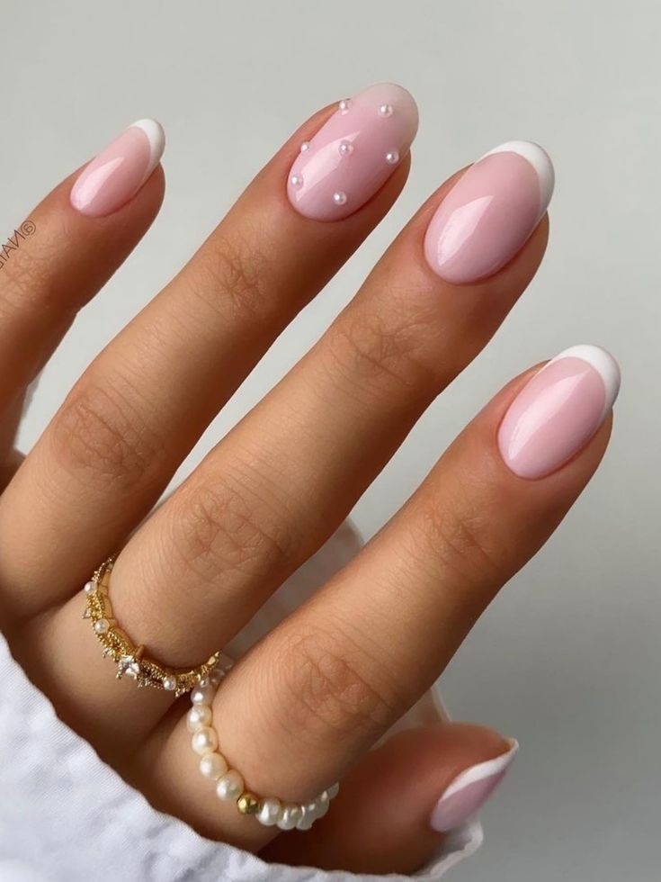 A hand featuring elegant, oval-shaped wedding day nails with a soft pink base and crisp white French tips, except for the index finger, which is adorned with scattered miniature pearls.