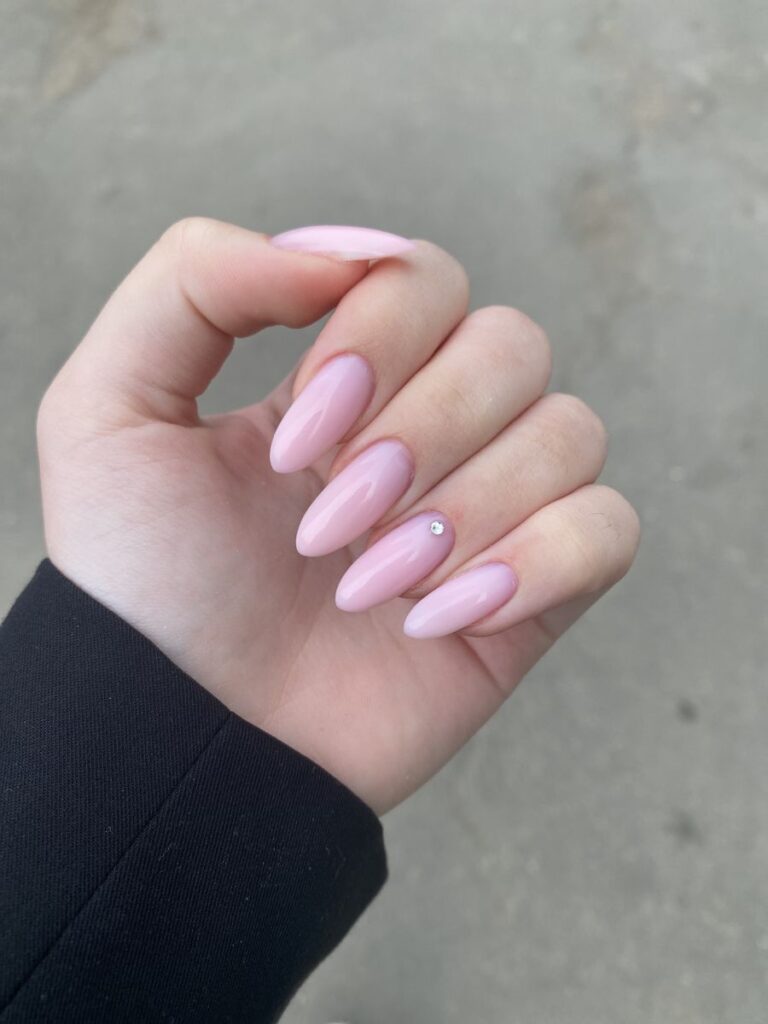 A close-up of a hand with flawless, elongated almond wedding nails in a glossy, opaque pastel pink, featuring a single small silver rhinestone placed at the base of the ring finger.