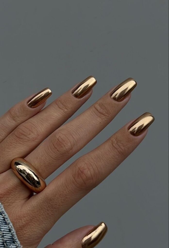 A close-up of a hand featuring medium-length square nails painted in a highly reflective, metallic liquid gold chrome finish. The person is wearing a chunky gold dome ring and a denim jacket.