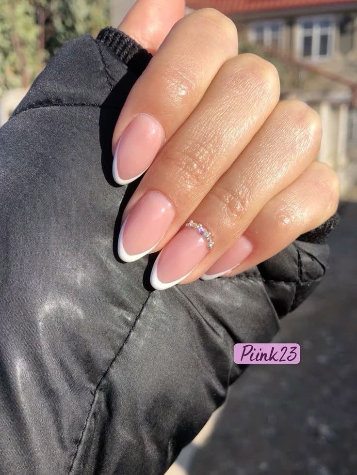 A hand clad in a black quilted jacket sleeve shows off a classic French manicure on long oval wedding nails, with a precise curve of micro-crystals framing the ring finger's cuticle.