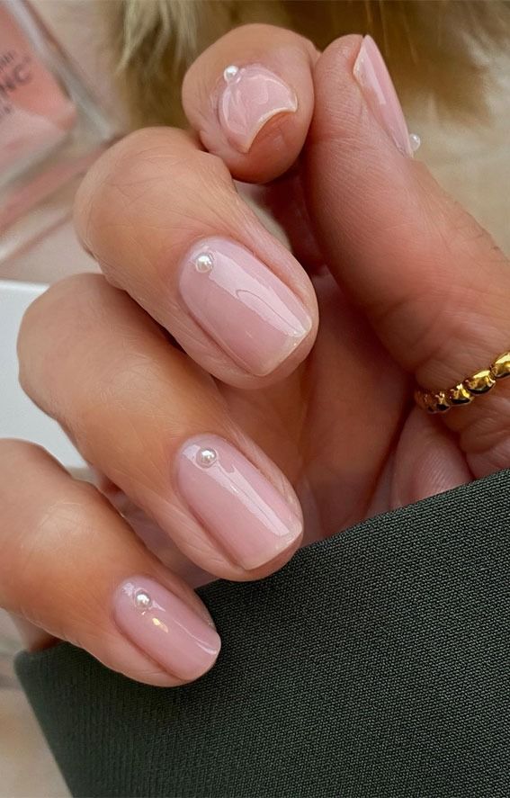 A close-up view against dark fabric and a bottle shows short, square-oval nails painted in a sheer nude, with a single, small pearl placed perfectly at the base of each bridal nail.