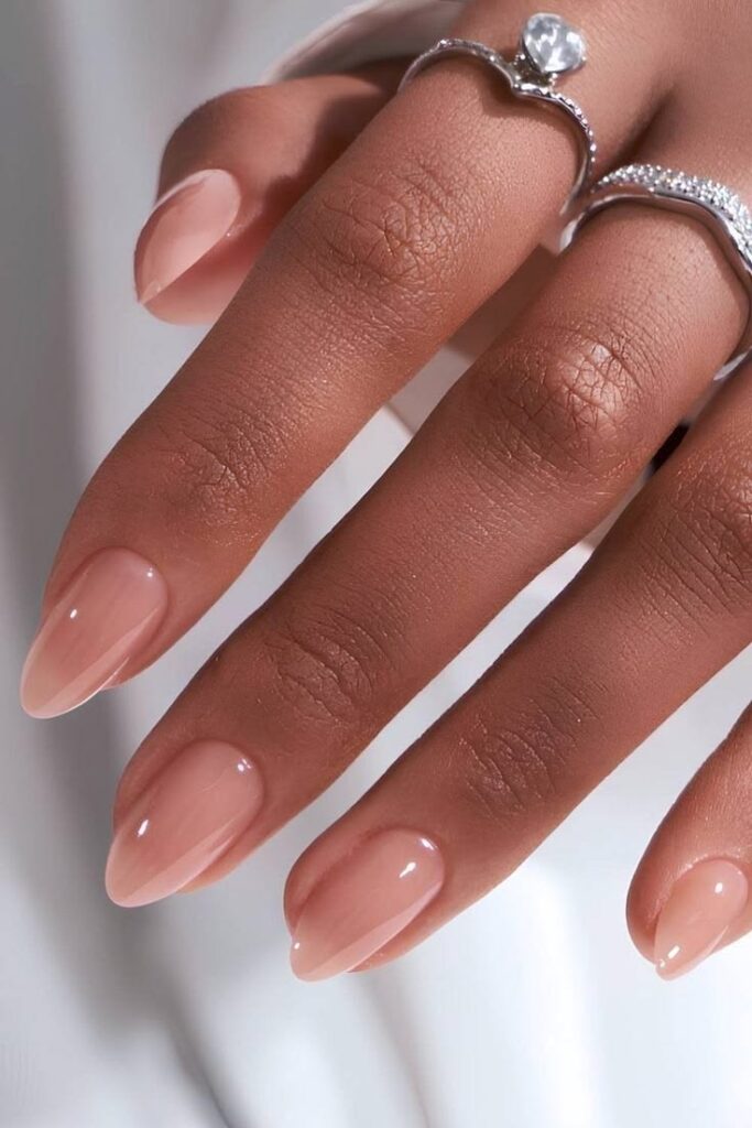 A high-end editorial photo of a hand with long, elegant almond-shaped wedding nails in a glossy, neutral peach-pink hue.