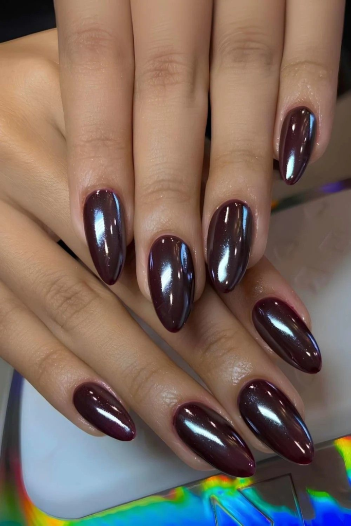 A top-down view of two hands crossed over, showing long almond nails coated in a deep, reflective dark purple and blackberry chrome finish with metallic silver highlights.