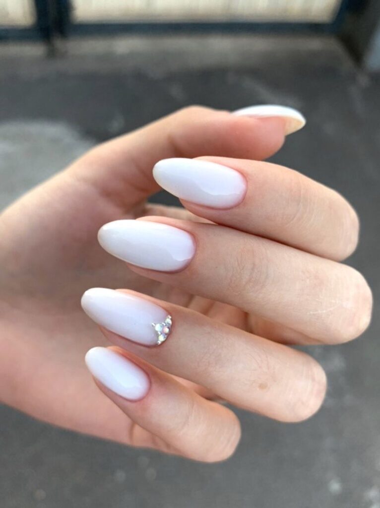 An outdoor shot of a hand with long, oval, milky white wedding day nails, accented by a tiny cluster of clear crystals at the base of the ring finger nail.