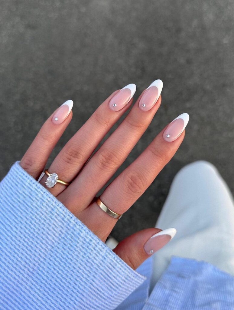An editorial-style photograph of a hand wearing an oval diamond engagement ring, displaying long, elegant French wedding nails accented by a single tiny rhinestone at the base of each finger.