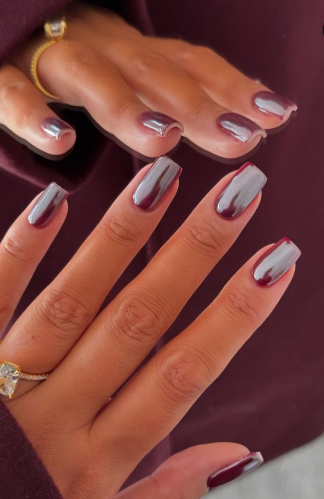 A hand with long square nails featuring a metallic plum-taupe chrome finish, reflecting soft purple and silver tones, with a prominent diamond ring visible on the finger.