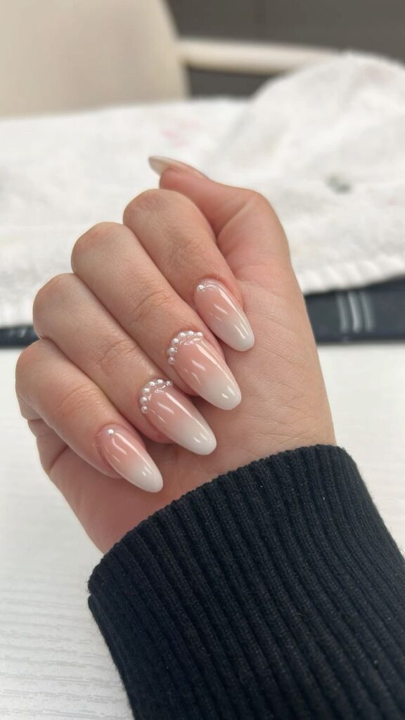 A hand wearing a black knit sleeve displays long, almond-shaped ombré bridal nails with arcs of tiny white pearls elegantly framing the cuticle lines.