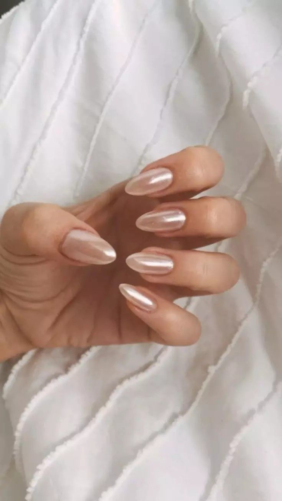 A hand with almond-shaped nails painted with a sheer, pearlized metallic polish against a white ruffled fabric.