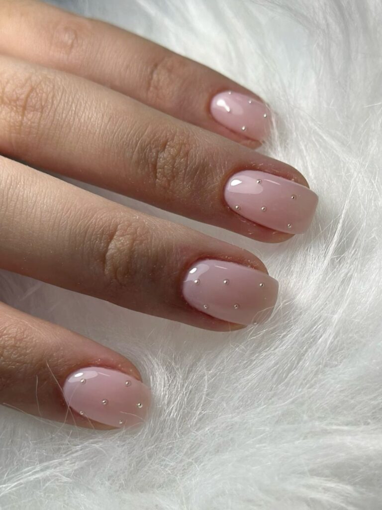 Short, neatly manicured nails against a white feathery background are polished in a glossy sheer nude and adorned with precisely placed silver microbeads for sophisticated nail ideas.