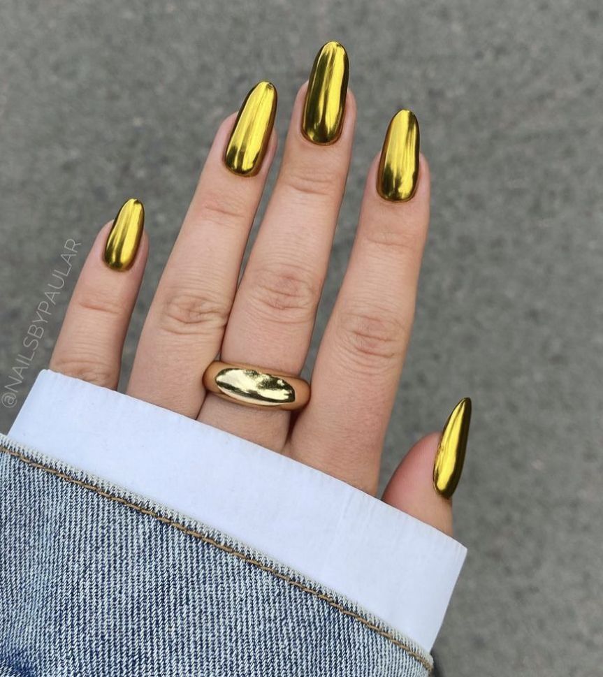 hand with long, almond-shaped nails coated in a brilliant, reflective chartreuse-gold chrome finish, accessorized with a thick gold ring against a soft gray background.
